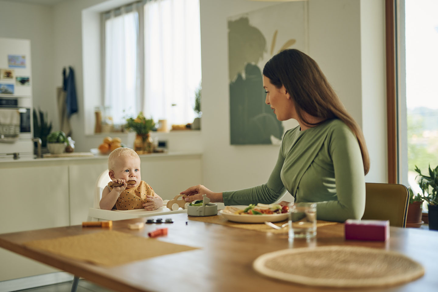 Diese Szene zeigt eine Mutter und ihr Baby, die gemeinsam an einem Tisch während der ersten Mahlzeit sitzen. Die Einführung von festen Nahrungsmitteln ist ein bedeutender Schritt in der Entwicklung des Babys. Mit einfachen, gesunden Mahlzeiten fördert die Mutter das Wohlbefinden und die gesunde Entwicklung des Kindes. Produkte wie Novalac unterstützen in dieser Übergangsphase, indem sie auf die speziellen Ernährungsbedürfnisse von Babys abgestimmt sind.
