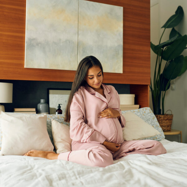 Eine schwangere Frau sitzt auf dem Bett und streichelt liebevoll ihren Bauch.