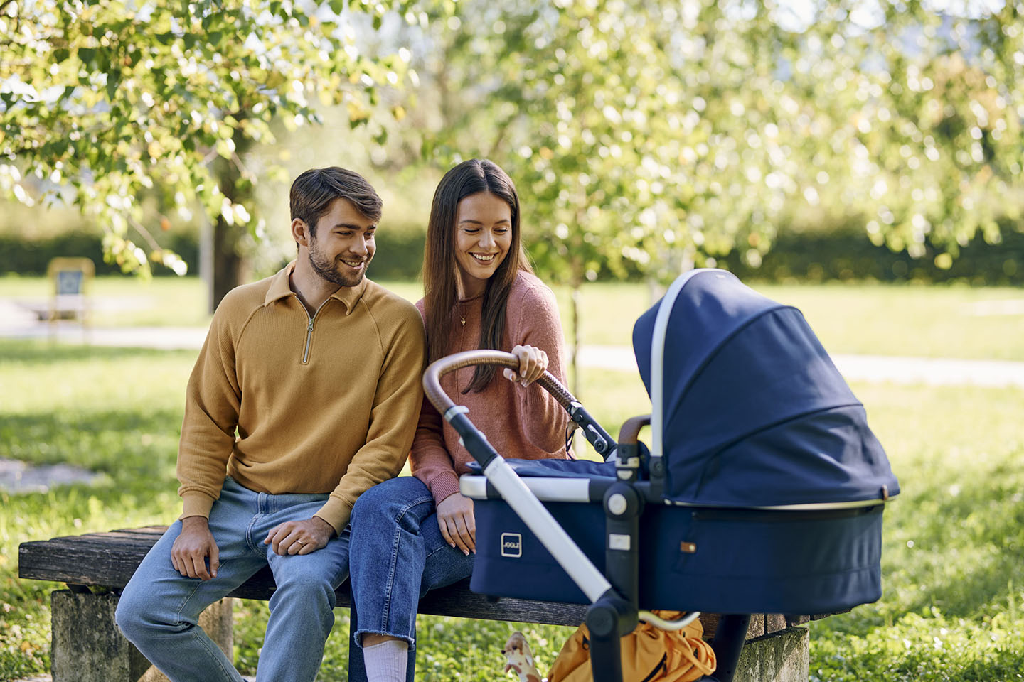 Diese Szene zeigt Eltern, die mit ihrem Kinderwagen auf einer Parkbank sitzen und einen Moment der Ruhe genießen. Solche Spaziergänge und gemeinsamen Pausen im Freien stärken die Familienbindung, fördern frische Luft und Entspannung und schaffen wertvolle Momente im Alltag mit einem Baby.