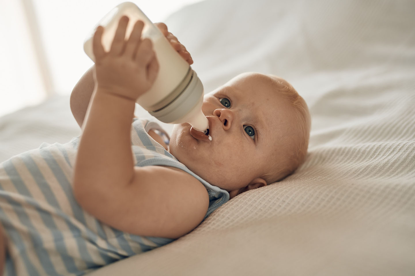 Diese Szene zeigt ein Baby, das auf dem Bett liegt und interessiert eine Novalac Milchflasche erkundet. Solche Momente fördern die natürliche Neugier und die motorische Entwicklung des Babys. Gleichzeitig verweist das Motiv auf die Bedeutung einer gut verträglichen und hochwertigen Säuglingsnahrung für das Wohlbefinden des Kindes.