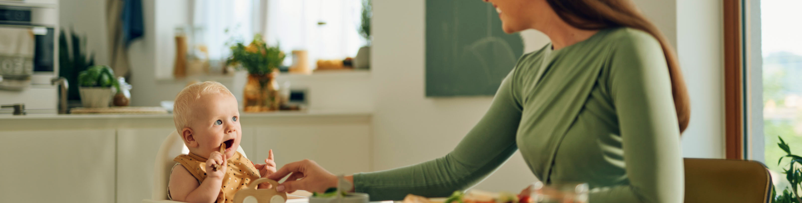 Diese Szene zeigt eine Mutter und ihr Baby, die zusammen am Tisch sitzen und eine Mahlzeit teilen. Die Einführung erster einfacher Beikost ist ein wichtiger Entwicklungsschritt für das Baby. Eine ruhige, positive Essenssituation unterstützt das Lernen neuer Geschmäcker und fördert gesunde Ernährungsgewohnheiten von Anfang an.