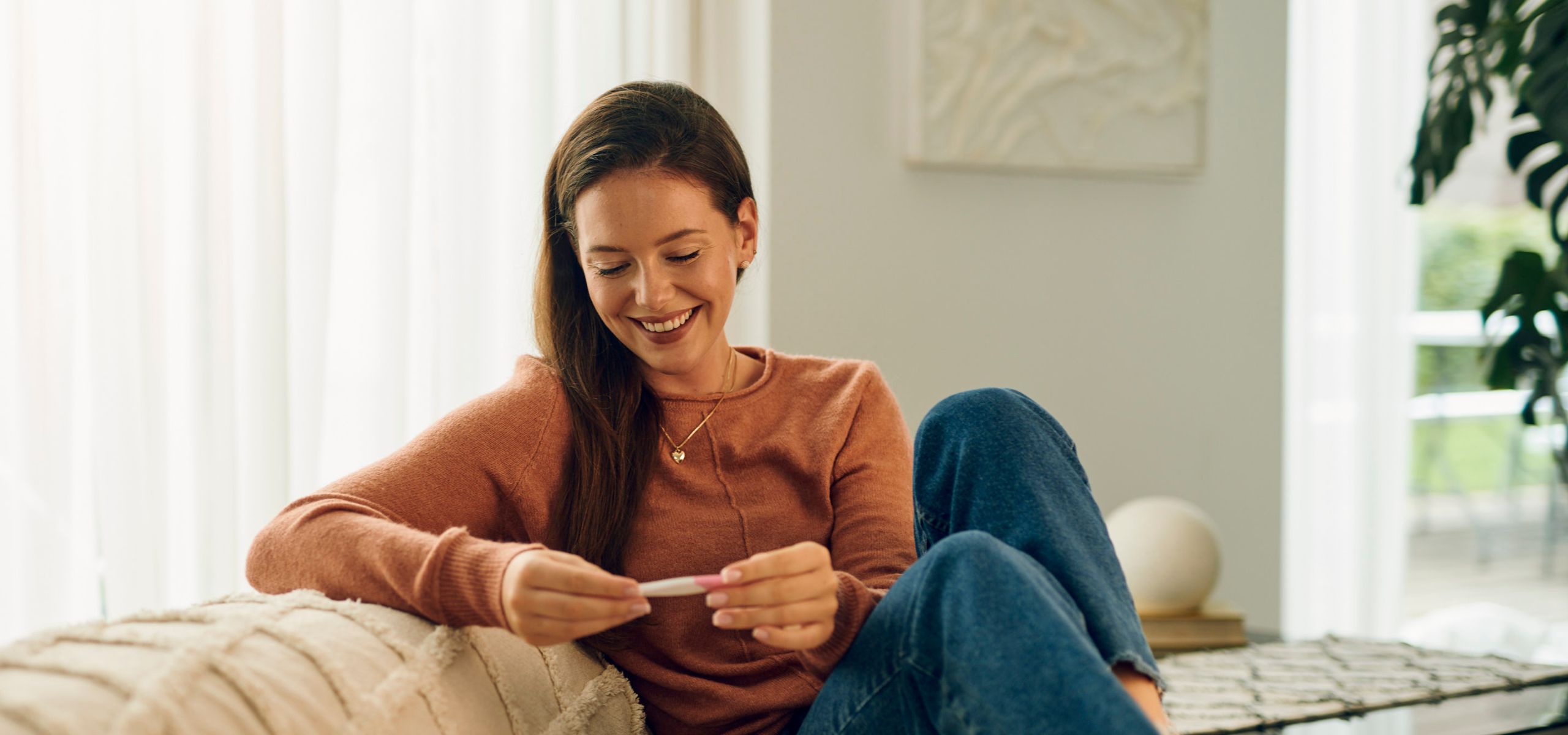 Eine schwangere Frau sitzt auf dem Sofa und schaut auf einen positiven Schwangerschaftstest, während sie ihre Schwangerschaft plant.