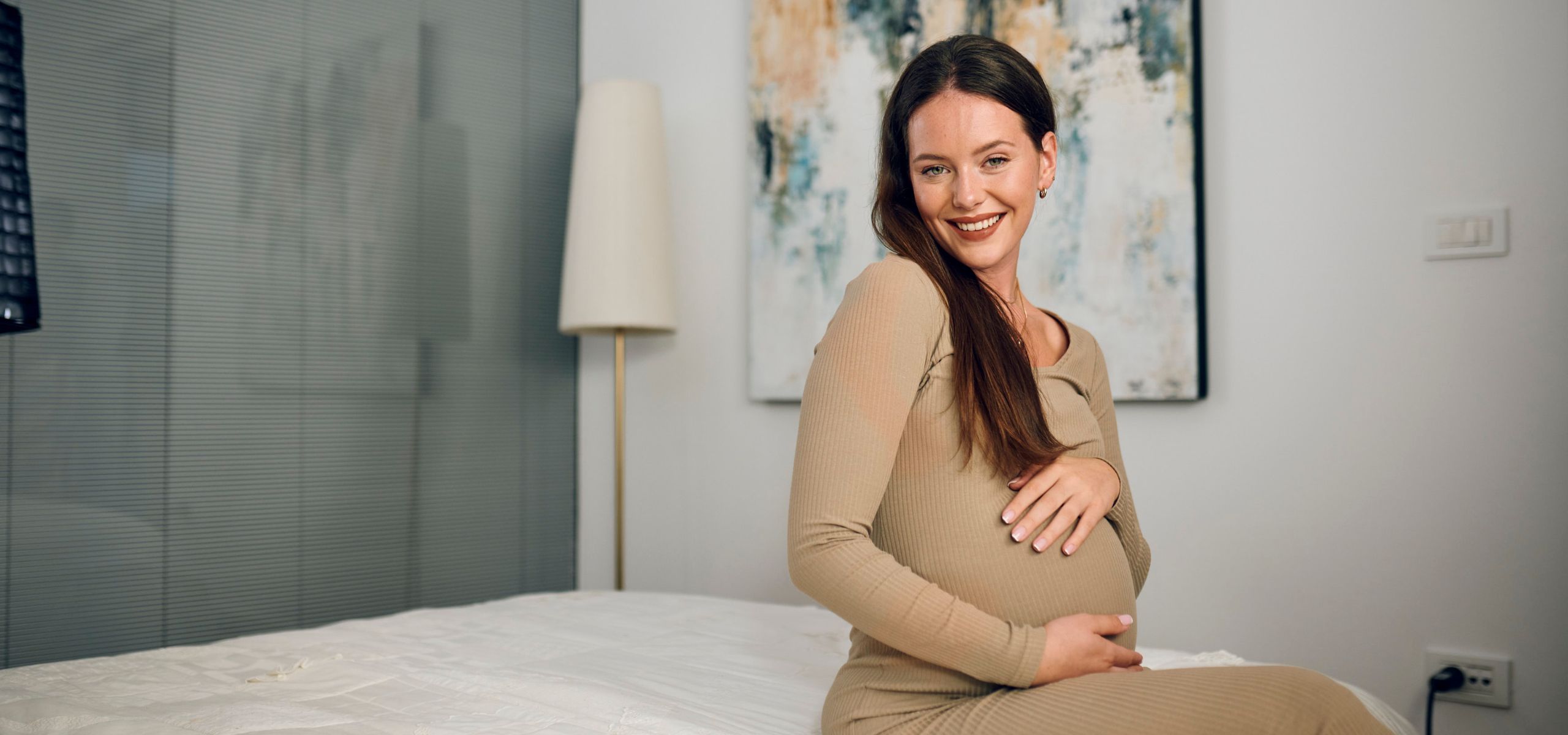 Eine schwangere Frau sitzt auf dem Bett und streichelt liebevoll ihren Bauch.