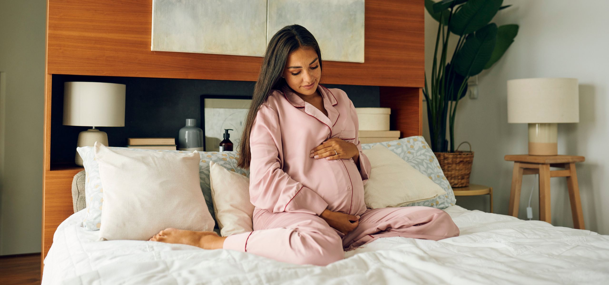 Eine schwangere Frau sitzt auf dem Bett und streichelt liebevoll ihren Bauch.