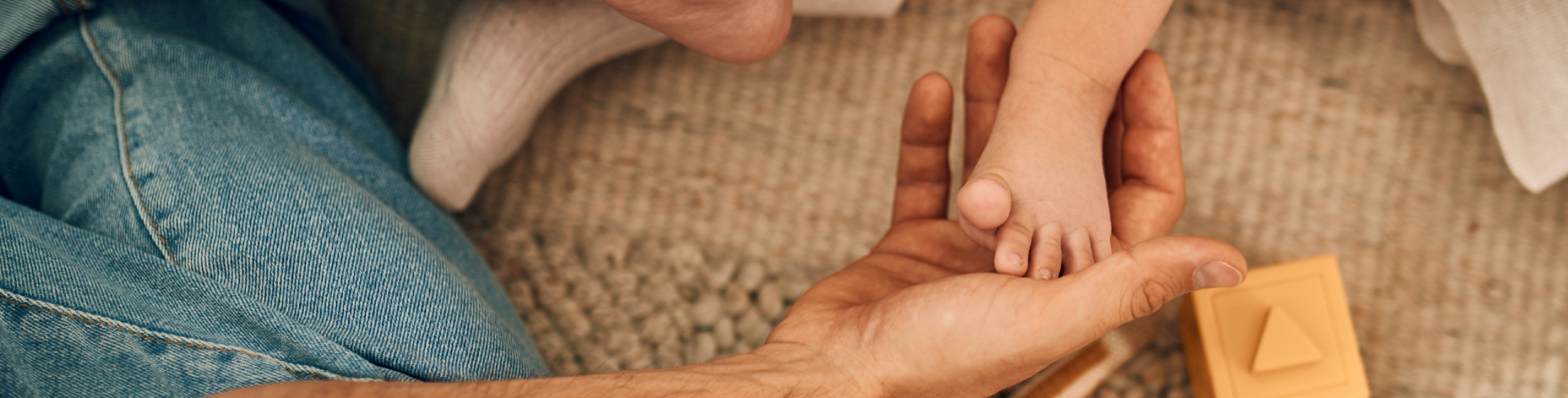Diese Szene zeigt einen Vater, der sanft den Fuß seines Babys hält, während das Neugeborene auf einem weichen Teppich mit Spielzeug liegt. Solche ruhigen, liebevollen Augenblicke stärken die Bindung und vermitteln dem Baby ein Gefühl von Sicherheit und Geborgenheit in seinen ersten Tagen zu Hause.