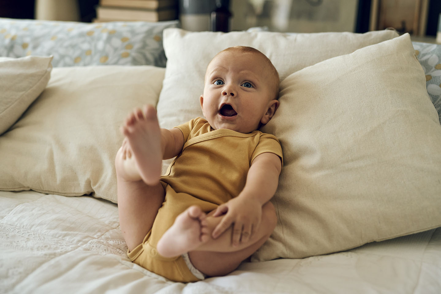 Diese Szene zeigt ein glückliches Baby, das entspannt auf Kissen liegt und spielerisch die Beine hebt. Solche Momente fördern die motorische Entwicklung, Bewegungsfreude und Wahrnehmung des eigenen Körpers. Die weiche Umgebung vermittelt Geborgenheit und schafft einen sicheren Raum für spielerisches Entdecken.