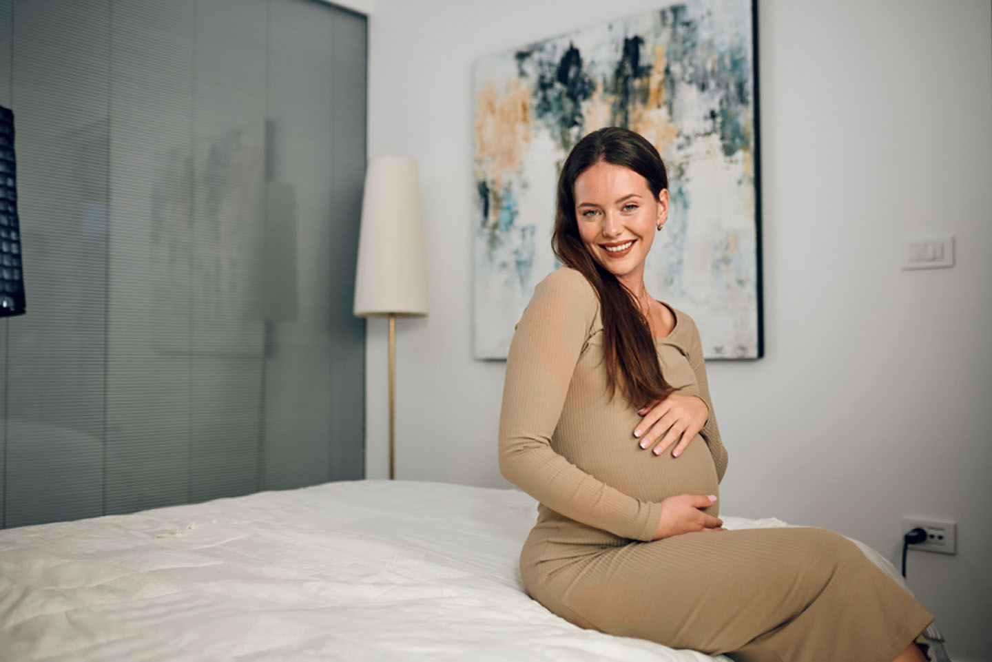 Diese Szene zeigt eine schwangere Frau, die auf dem Bett sitzt und lächelnd in die Zukunft blickt. Der Moment vermittelt Ruhe, Wohlbefinden und Vorfreude auf das kommende Baby. Solche positiven Augenblicke stärken das emotionale Gleichgewicht und unterstützen eine gesunde Schwangerschaft.